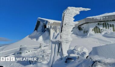 Bright blue skies are the back drop to a building with snow up the sides and on the ground and fence all around.