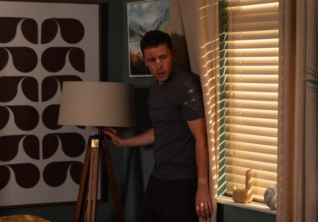 Todd Grimshaw hiding behind a curtain in his flat in Coronation Street.