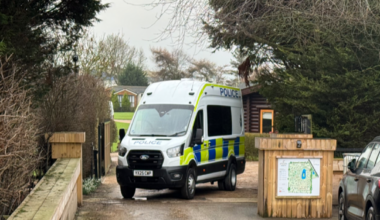 The picture shows a police van which has been parked across an entrance. There is a wooden block with a map on it to the right-hand side of the van. Behind it is a wooden log cabin.