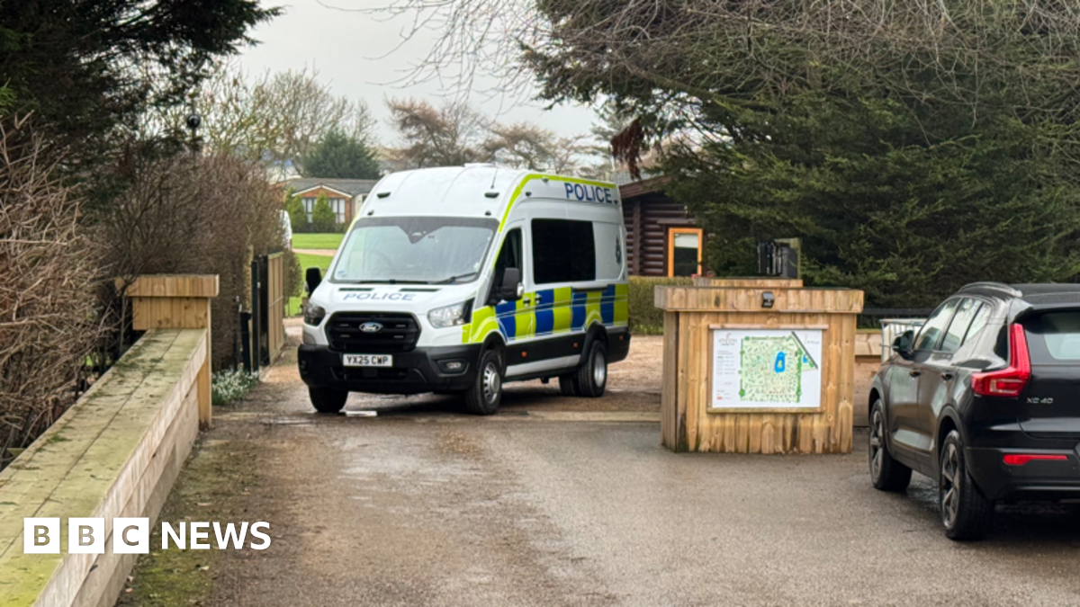 The picture shows a police van which has been parked across an entrance. There is a wooden block with a map on it to the right-hand side of the van. Behind it is a wooden log cabin.