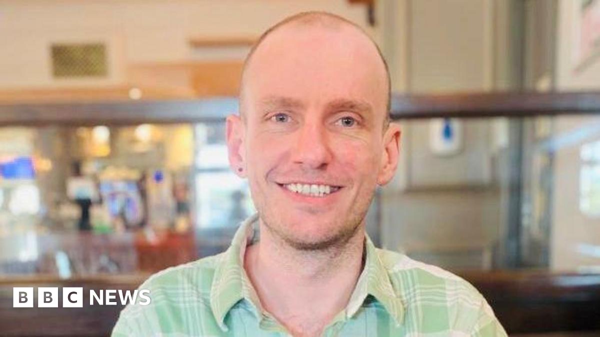 Ben Howard, a man with a buzz cut and stubble, is pictured smiling at the camera. He is wearing a light green, checkered shirt with a collar. He appears to be sat in a pub.