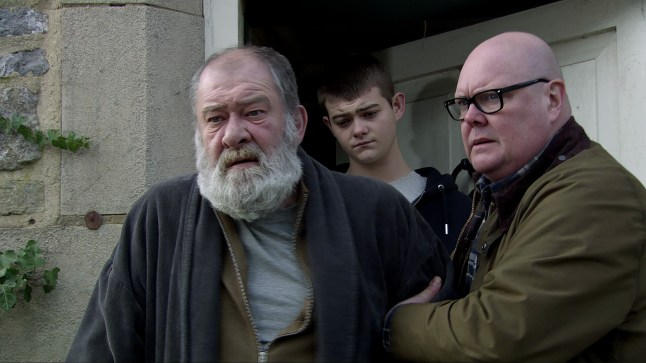 Paddy Kirk and Dylan Penders trying to help a distressed Bear Wolf in Emmerdale, as they lead him out of a house front door.