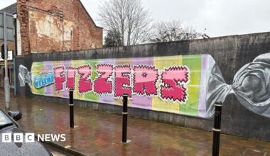 A long wall beside a wet sidewalk shows a mural painted like a wrapped candy. It has pastel stripes and large pink letters spelling “FIZZERS.”