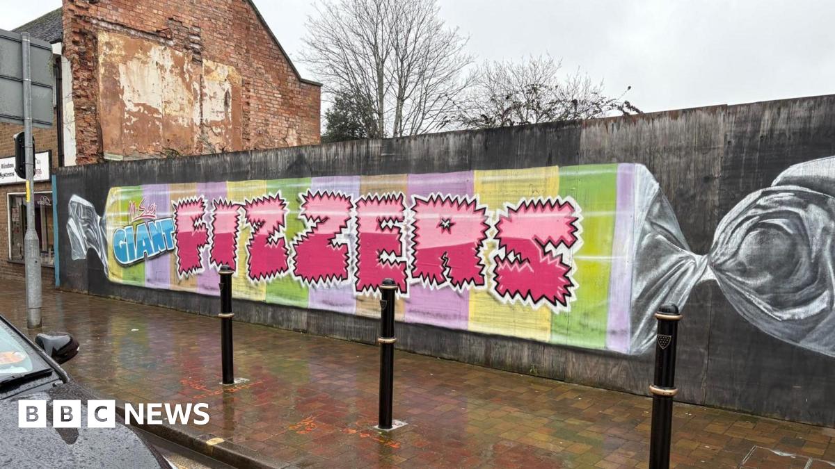 A long wall beside a wet sidewalk shows a mural painted like a wrapped candy. It has pastel stripes and large pink letters spelling “FIZZERS.”