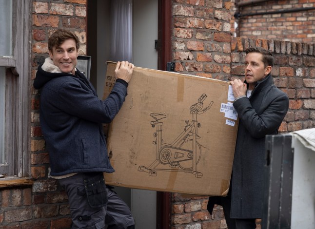 Coronation Street's Theo Silverton and Todd Grimshaw stood outside their flat holding a large box containing an exercise bike.