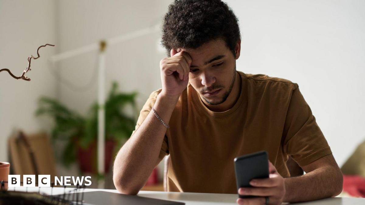 A young man looks at his phone while sitting at a computer in his home. He looks weary.