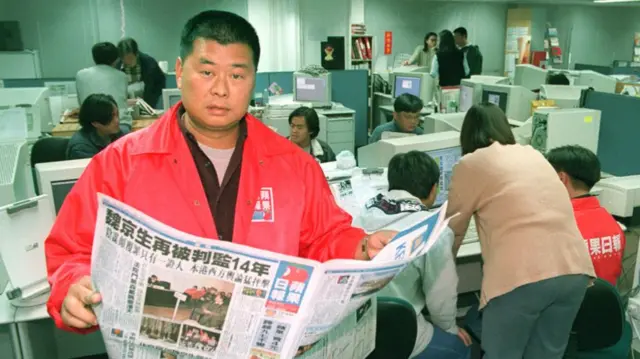 Jimmy Lai in a red jacket standing in a newsroom, holding a newspaper