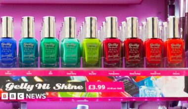 Man with dark hair and brown eyes, wearing a mid-blue button-up shirt, sits at a table on which there is an array of nail vanishes in different colours including pink, purple, lilac, green, yellow and orange polishes.