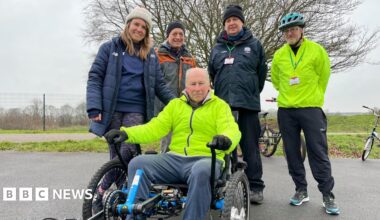 A man sits in a wheelchair trike wearing a high vis jacket. Around him stand 4 people smiling from the track. To the left is a young lady who wears a white hat and blue coat, then an older man who wears a black hat and light grey coat with orange detailing, next stands a man in another black hat wearing a dark grey coat and then stands a man in a high vis jacket and helmet.
