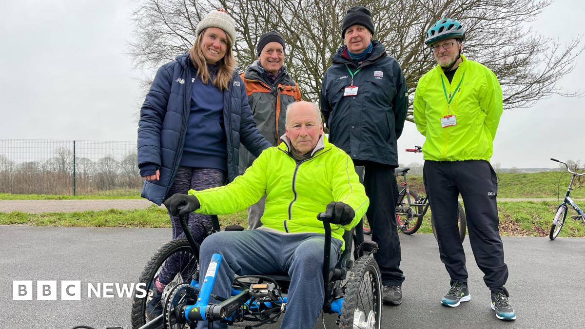 A man sits in a wheelchair trike wearing a high vis jacket. Around him stand 4 people smiling from the track. To the left is a young lady who wears a white hat and blue coat, then an older man who wears a black hat and light grey coat with orange detailing, next stands a man in another black hat wearing a dark grey coat and then stands a man in a high vis jacket and helmet.