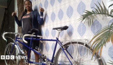 A woman wearing a black T-shirt and blue jacket smiles and holds two thumbs up  while standing behind a blue tinted tandem bike