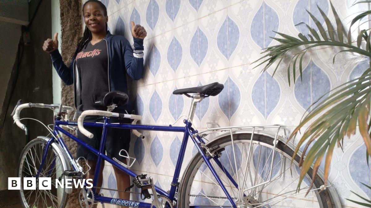 A woman wearing a black T-shirt and blue jacket smiles and holds two thumbs up  while standing behind a blue tinted tandem bike