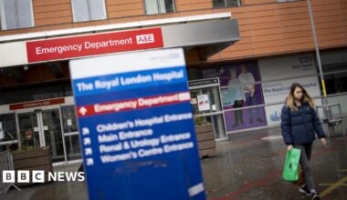 A view of the front entrance to the Royal London Hospital's emergency department on a wet day. The building is clad is long brown tiles, with a protruding steel eave which reads: Emergency Department A&E. A woman carrying a green plastic bag is on the right of the image, and blue hospital signage is out of focus in the image foreground, which reads: The Royal London Hospital.