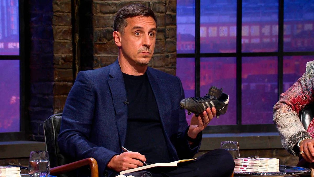 An individual dressed in a blue suit is seated in a chair, holding a pair of shoes, and appears to be engaged in a conversation.