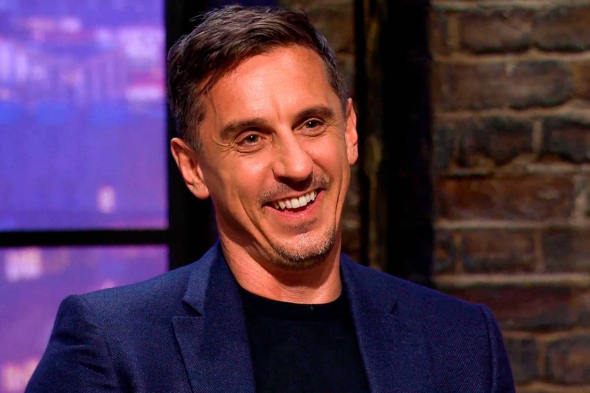 An individual, dressed in a navy blue suit and a black inner shirt, is seated and smiling brightly in front of a brick wall backdrop.