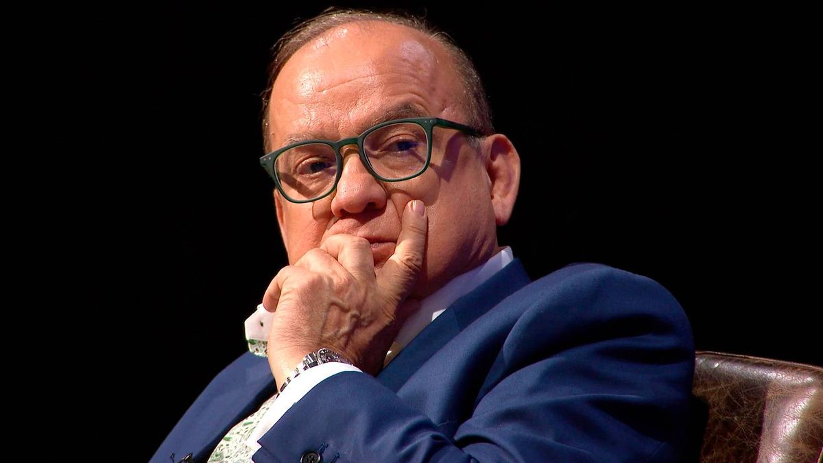 An individual dressed in a dark blue suit, complete with a white shirt and black-framed glasses, is seated and appears to be in deep thought, with one hand resting on his chin.