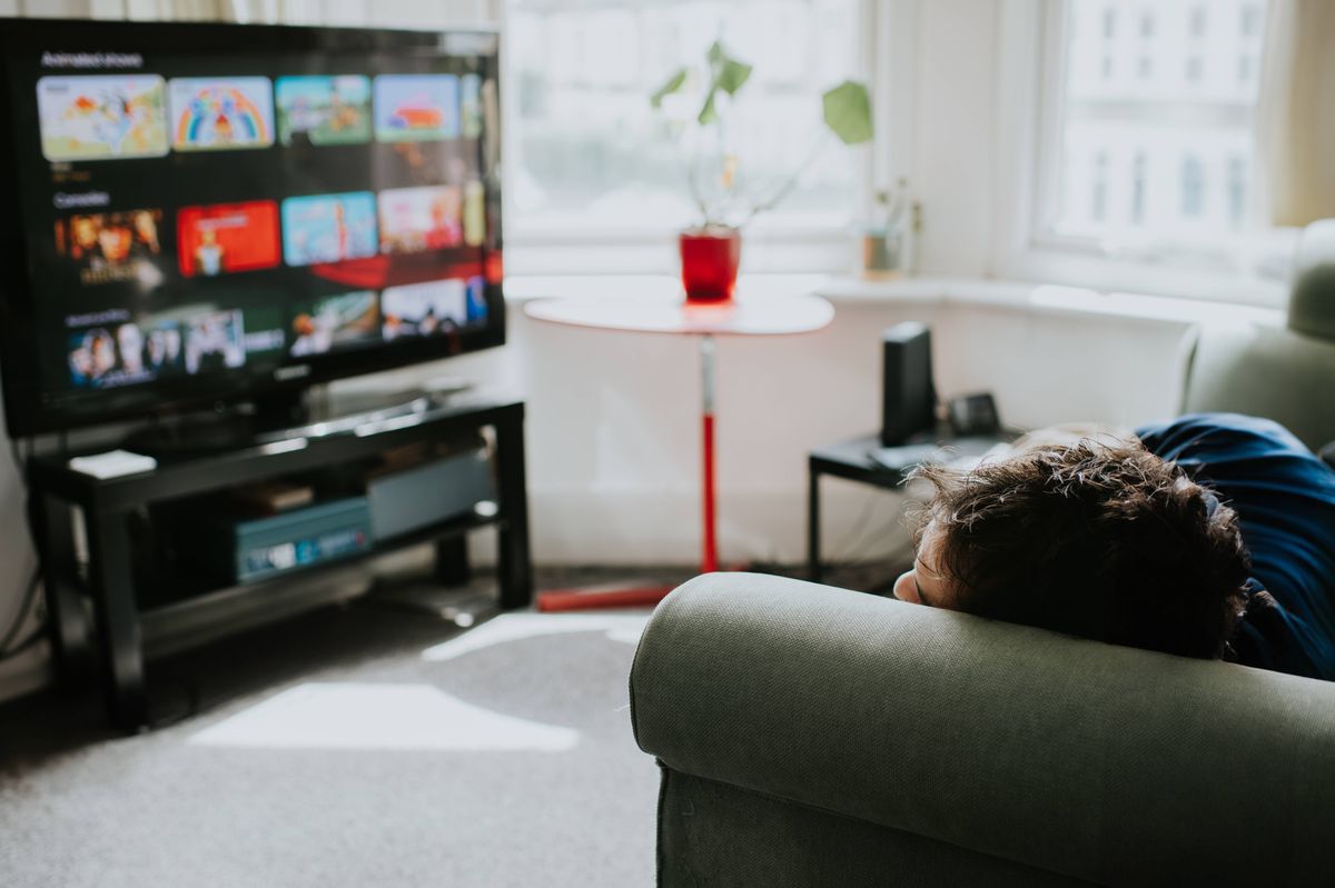A person relaxes on a comfortable couch choosing a programme on a streaming service. The warm, cozy living space features natural light streaming through the windows, creating a tranquil environment perfect for unwinding and enjoying entertainment.