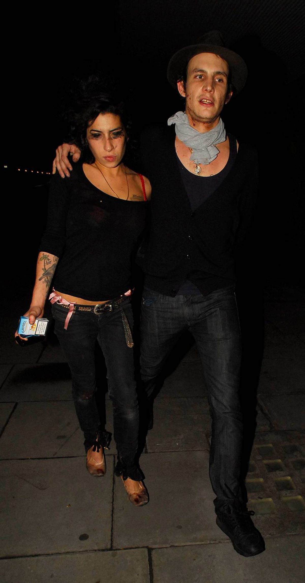 Two individuals, a man and a woman, are standing closely together, perhaps posing for a photograph in a dimly lit environment. The man is wearing a gray scarf around his neck, and both individuals are dressed in black clothing.