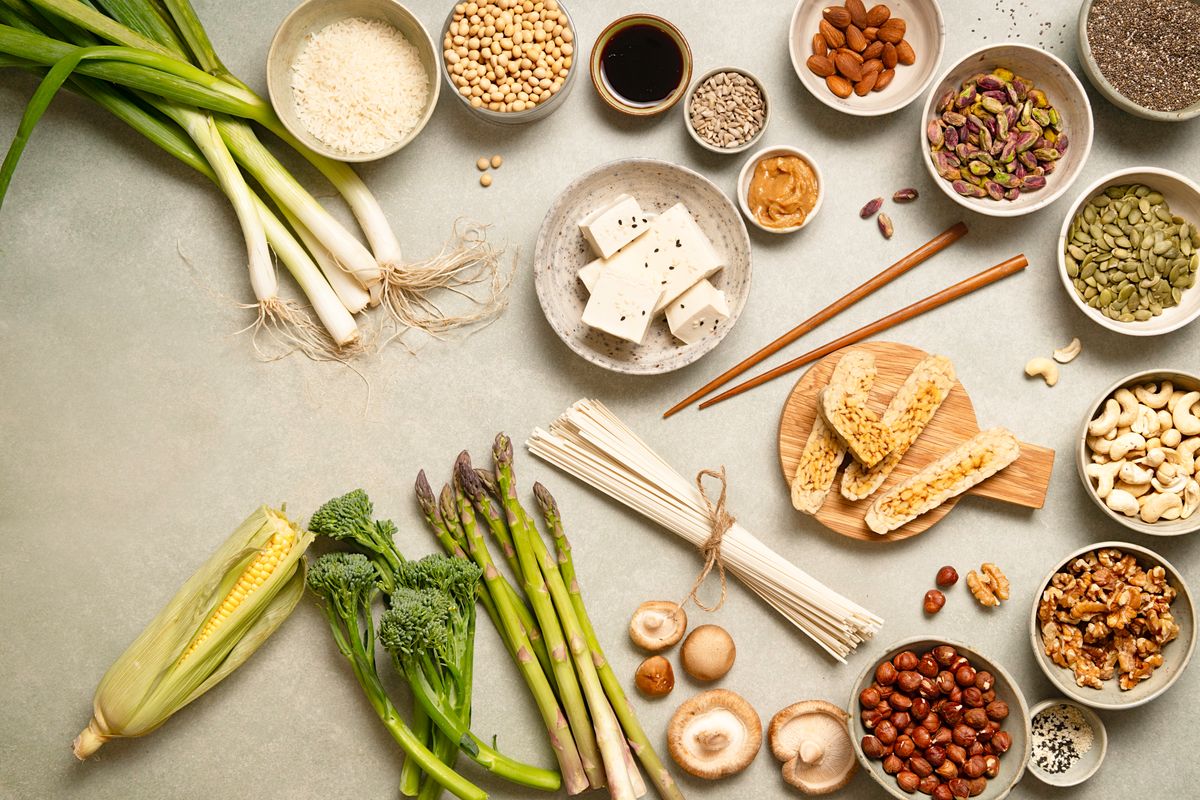 raw plant based and protein products on table in bowls and plates, soy beans, tofu, tempeh, almonds, macadamia, pistachio, chia seeds, nuts, soba, rice, miso, various types of seeds, fresh veggies, like corn, broccolini, spring onions, asparagus, directly above picture with copy space