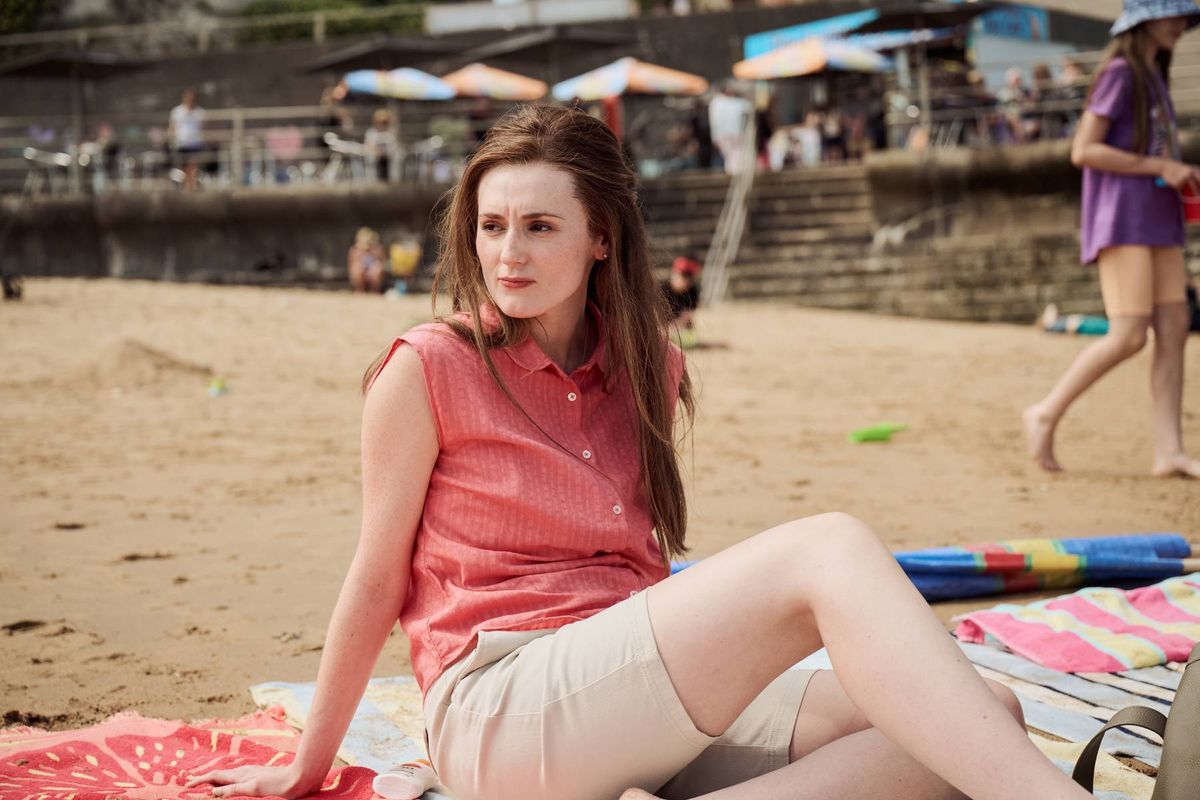 A woman is seated on a sandy beach, dressed in a sleeveless top and shorts, and appears to be resting on a blanket. In the background, another individual is seen walking along the shore. Various beach umbrellas and an elevated structure are also visible in the scene.