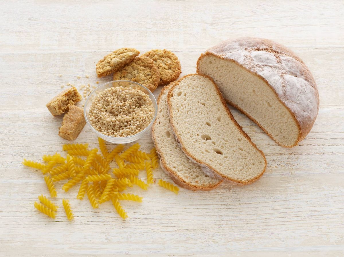 A collection of various food items is displayed on a white surface. These items include slices of bread, pasta, and a bowl of a grain-based substance. The presentation highlights the diverse textures and forms of the food, showcasing an assortment of whole grains and carbohydrates.