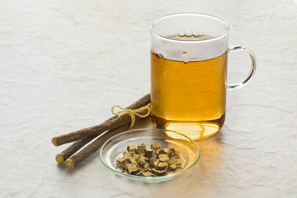 A clear glass cup filled with a light yellow liquid, possibly an herbal infusion, accompanied by a small glass bowl containing various small, dried herbs, and a bundle of cinnamon sticks on a white surface.