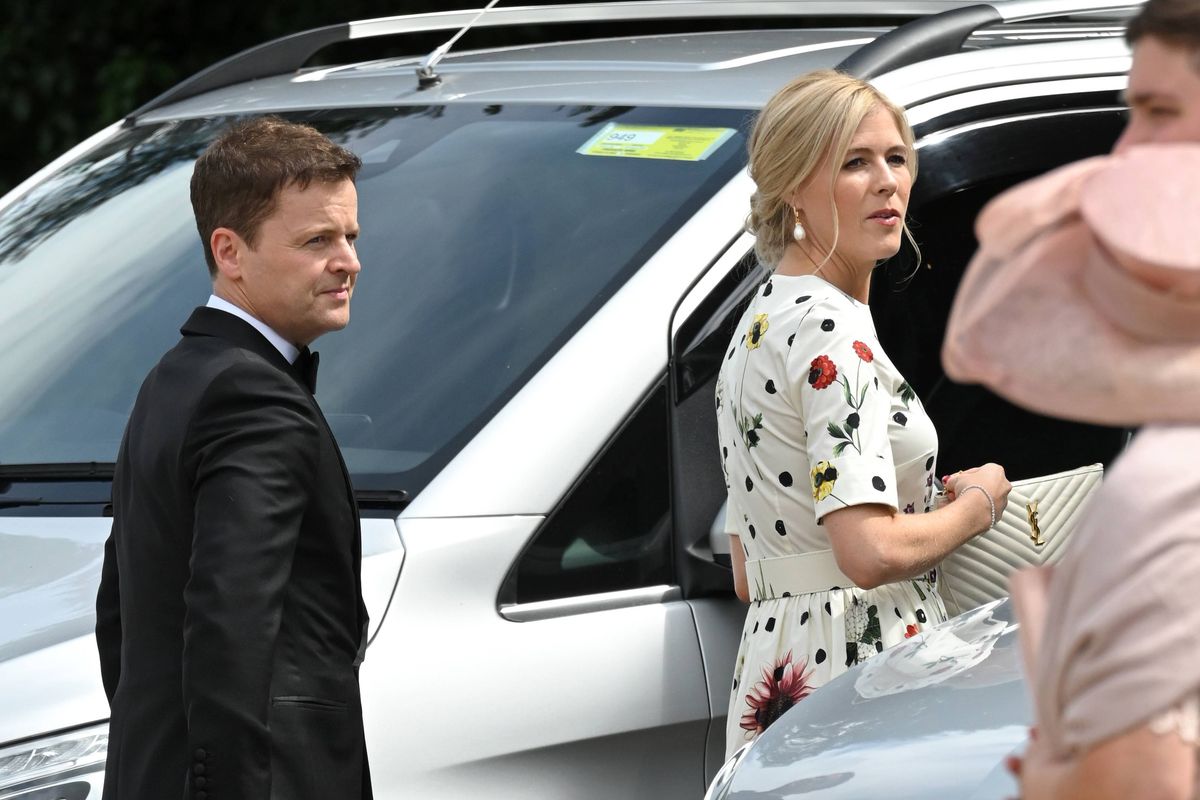 An individual in formal attire stands near a white car while another individual in a white floral dress converses with someone partially visible, creating a scene of engagement and interaction.