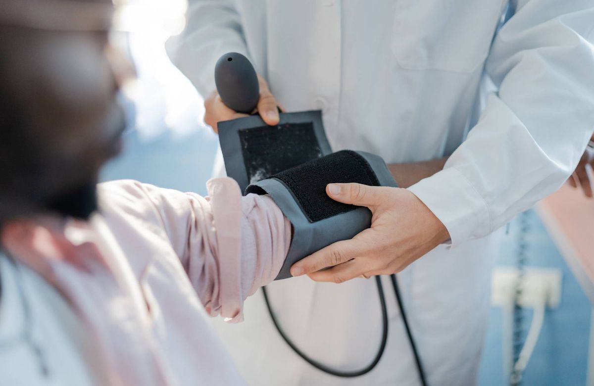 An individual in a white coat, presumably a healthcare professional, is using a sphygmomanometer to measure the blood pressure of another individual, who is dressed in a light-colored shirt. The setting appears to be a clinical or medical environment.