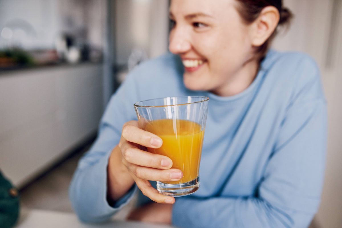 A cheerful woman in a light blue shirt holds a glass of orange juice, smiling and looking away