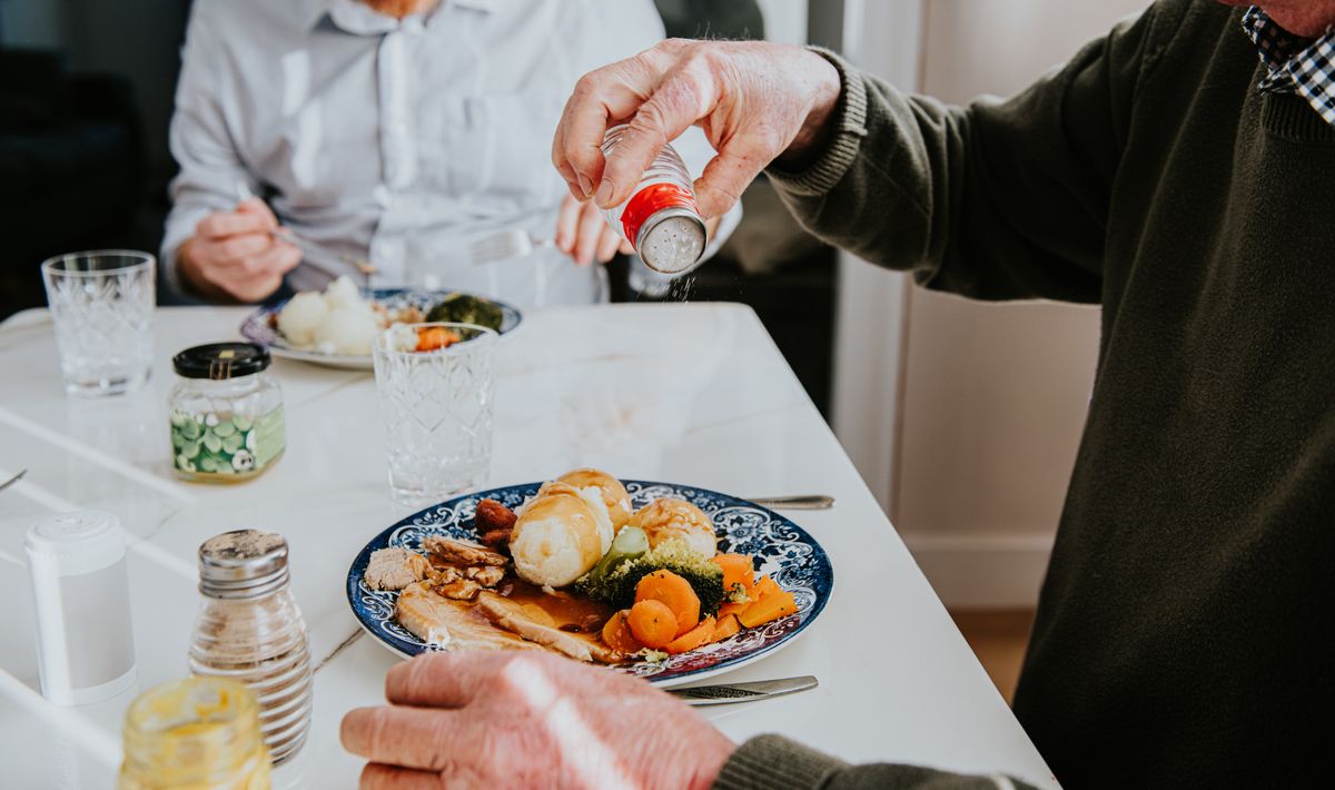 A man pours salt onto his plate filled with vegetables and meat while dining with another individual in a warm, inviting room, creating a comforting atmosphere.