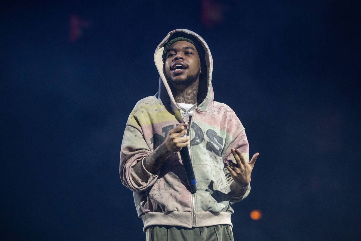 An individual dressed in a light-colored hoodie with a hood raised, standing on a stage and gesturing with both hands, possibly during a performance. The background is predominantly blue with spotlights illuminating the figure.