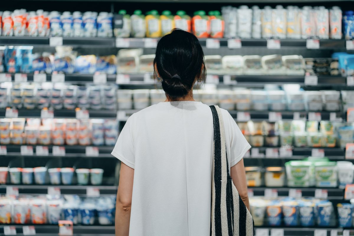Rear view of young Asian woman doing grocery shopping, standing in front of a shelf full of fresh dairy products in the dairy aisle and thinking which product to choose from in a supermarket.
