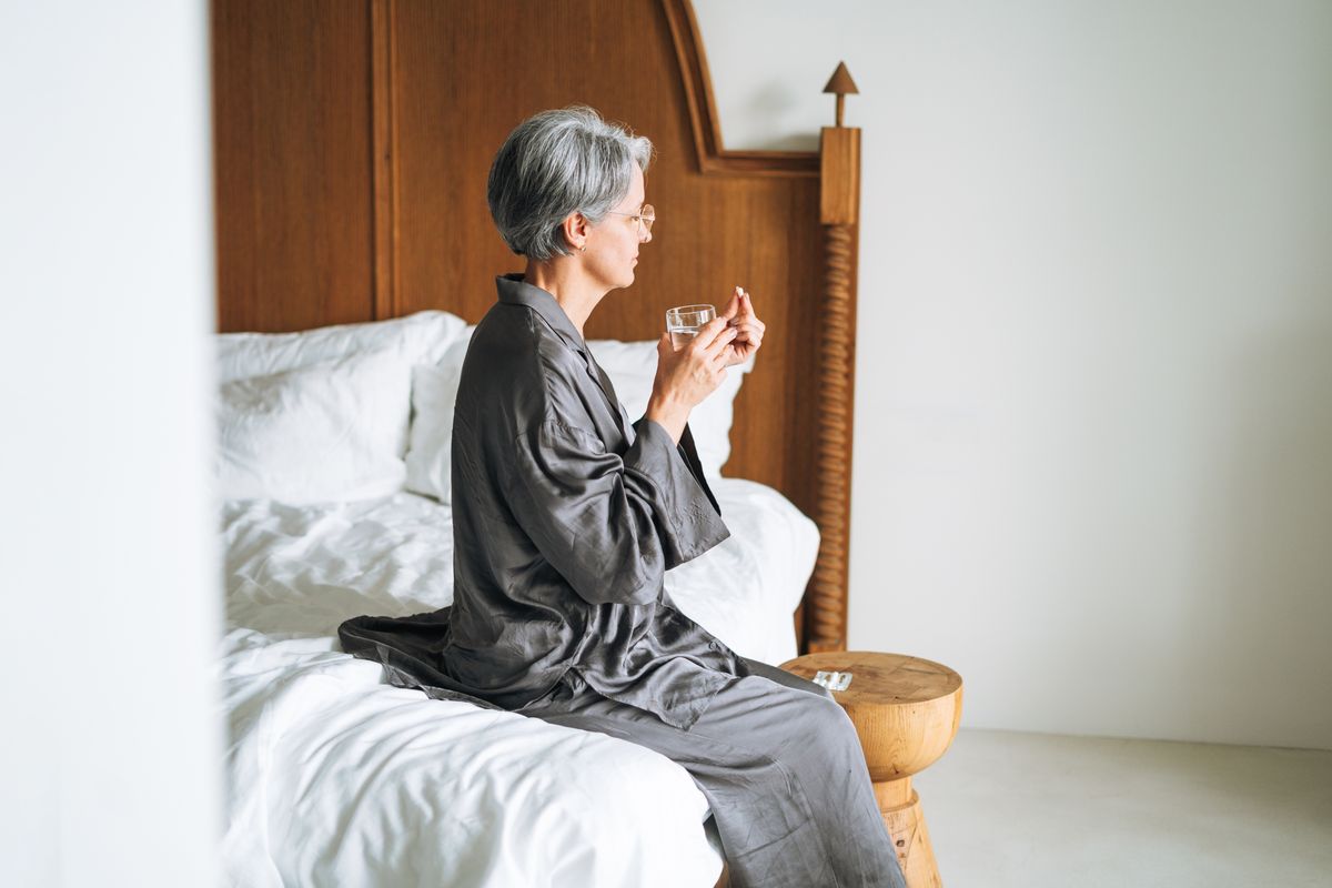 A elderly woman with gray hair drinks pills while sitting on her bed at home