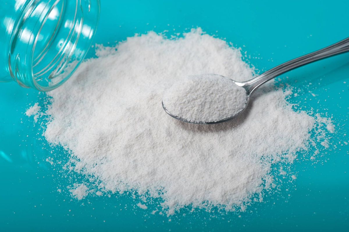 A heap of granulated sugar is depicted on a blue surface, with a silver spoon resting on top. A glass container, likely partially filled with sugar, is positioned to the left of the sugar pile.
