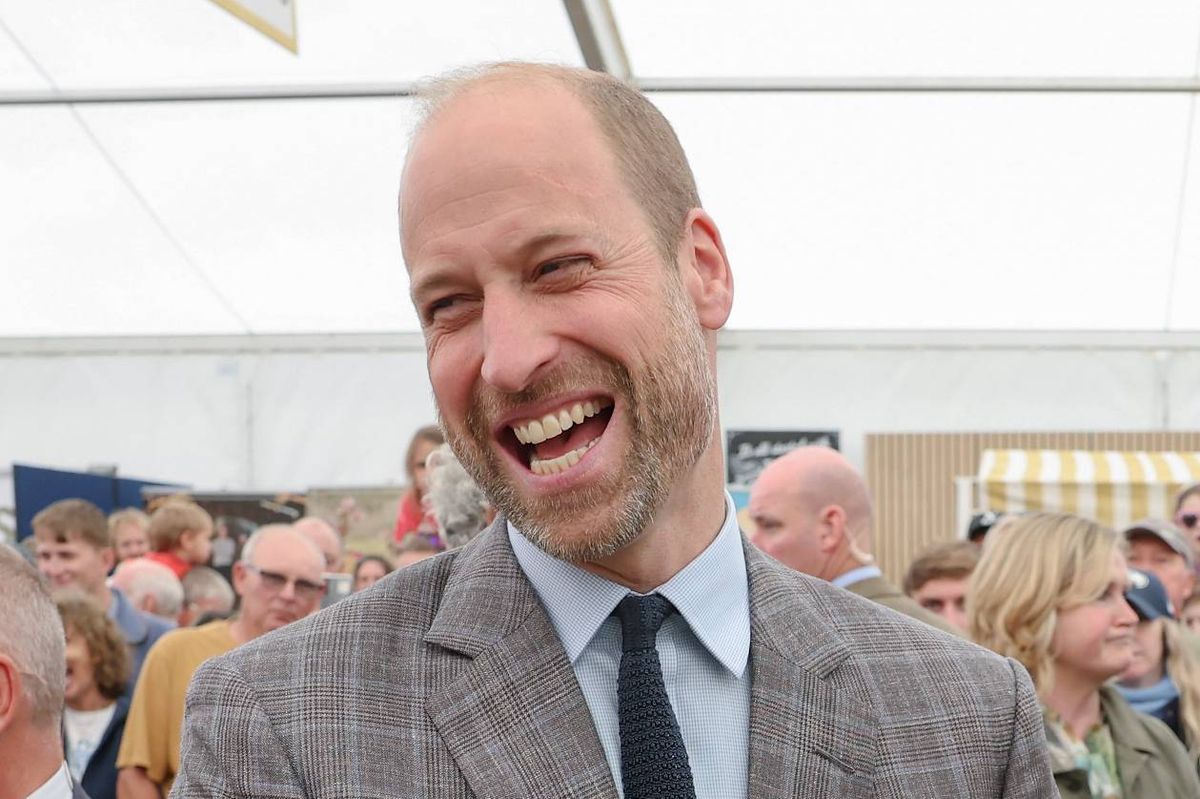 An individual in formal attire, consisting of a gray suit, a white shirt, and a black tie, is standing in a public setting and is visibly engaged in laughter. The background shows a blurred crowd of people, some of whom are also smiling, suggesting a festive or celebratory atmosphere. The setting appears to be an indoor event with a white ceiling and ambient lighting.