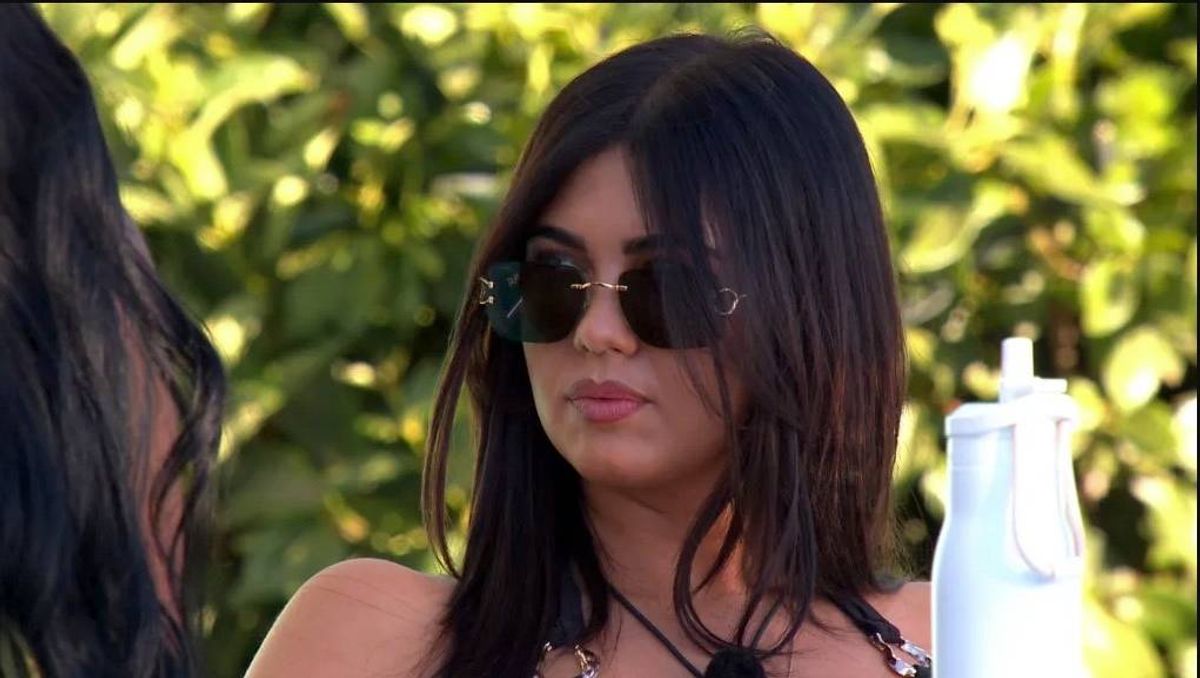 An individual with long, dark hair, donning sunglasses, stands in an outdoor setting with foliage in the background. The person is holding a white bottle in one hand.