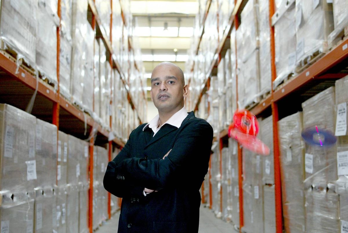 An individual in a formal suit stands confidently within a large warehouse, with rows of neatly organized shelves and various storage boxes in the background.