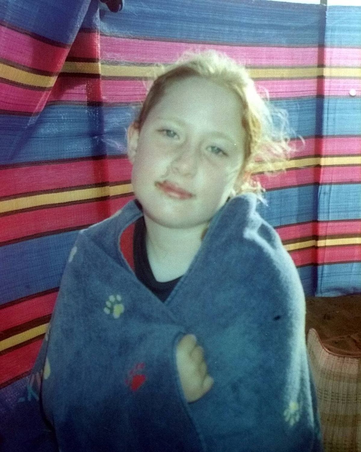 An image featuring a young individual with light hair, wearing a blue jacket, standing against a backdrop of vibrant, multicolored stripes. The individual is gazing directly at the camera, with a neutral expression.