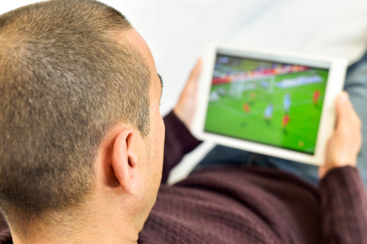 a young man lying on the couch watches football on a tablet.