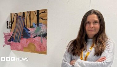 Belinda Downing is standing next to her artwork. She is wearing a grey jumper and is looking directly at the camera with her arms crossed.