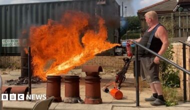 A street scene involving several vehicles and military personnel. In the foreground on the left, there is a white van with bright yellow and red diagonal stripes on the rear and text that reads “MOBILE CCTV UNIT Safer Neighbourhood Service.” To the right, there is a blue van parked nearby. In the centre of the image is a large white vehicle with open rear doors. Two individuals in camouflage uniforms are standing near the open doors, handling equipment, including a blue container on the ground and various gear inside the vehicle. The background shows brick buildings and greenery.
