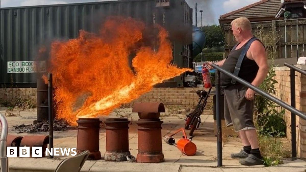A street scene involving several vehicles and military personnel. In the foreground on the left, there is a white van with bright yellow and red diagonal stripes on the rear and text that reads “MOBILE CCTV UNIT Safer Neighbourhood Service.” To the right, there is a blue van parked nearby. In the centre of the image is a large white vehicle with open rear doors. Two individuals in camouflage uniforms are standing near the open doors, handling equipment, including a blue container on the ground and various gear inside the vehicle. The background shows brick buildings and greenery.