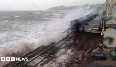 A damaged section of the coastal railway at Dawlish in 2014, with waves washing over broken track where the sea wall has collapsed. Houses sit close to the line on the right under a grey sky.