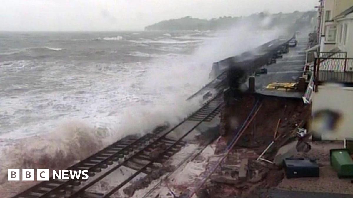 A damaged section of the coastal railway at Dawlish in 2014, with waves washing over broken track where the sea wall has collapsed. Houses sit close to the line on the right under a grey sky.