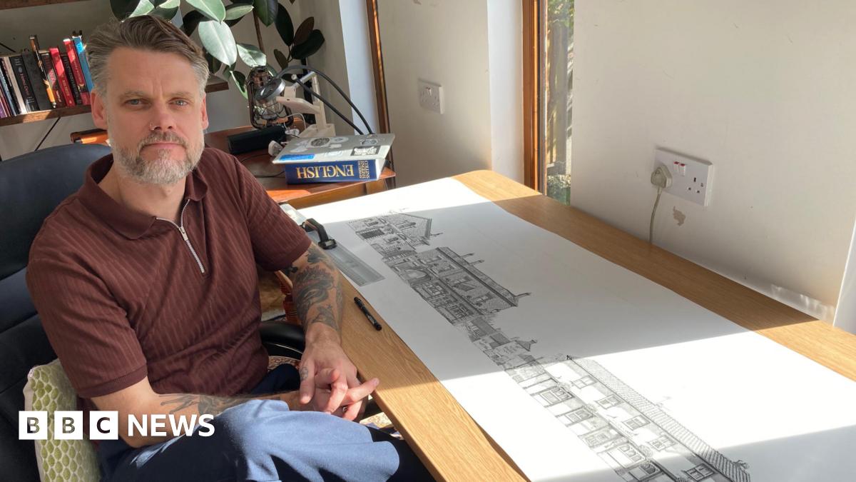 A man with grey and brown hair slicked away from his forehead is sat in a chair next to a desk looking at the camera. He has his hands together and knee crossed and is wearing a brown short-sleeved shirt and blue trousers. On the table is a long paper roll with detailed drawings of buildings.