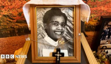 An old black and white photo of May wearing her uniform. The photo is in a wooden frame. A necklace with a cross on it is attached to the frame. Fabric is draped over the frame which sits on a mantlepiece.