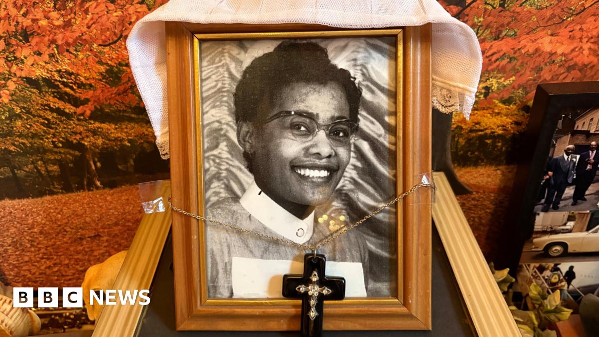 An old black and white photo of May wearing her uniform. The photo is in a wooden frame. A necklace with a cross on it is attached to the frame. Fabric is draped over the frame which sits on a mantlepiece.