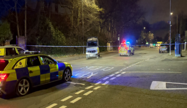 The scene of a fatal collision on a road at night, the area is cordoned off with police tape and there are several police cars at the scene with flashing lights.