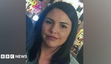 Alice Webb is smiling at the camera, she has dark hair and a piercing in her lower lip. She stands in front of an illuminated bar.