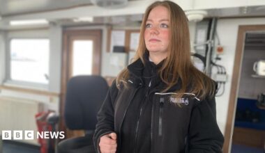 Ellie stands behind the wheel of the ferry, looking out to sea. She has brown hair and blue eyes and is wearing a black zip-up fleece.
